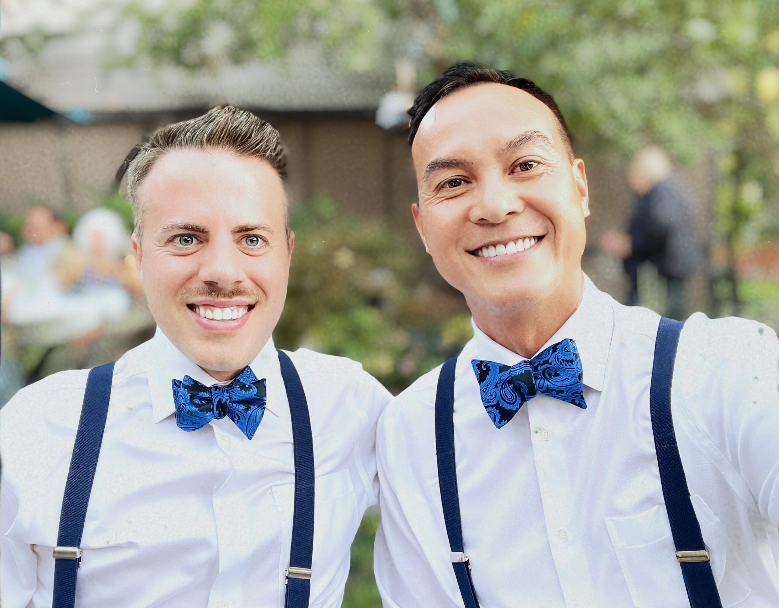 The couple smiling at a garden party. They both are wearing blue bow ties and suspenders.